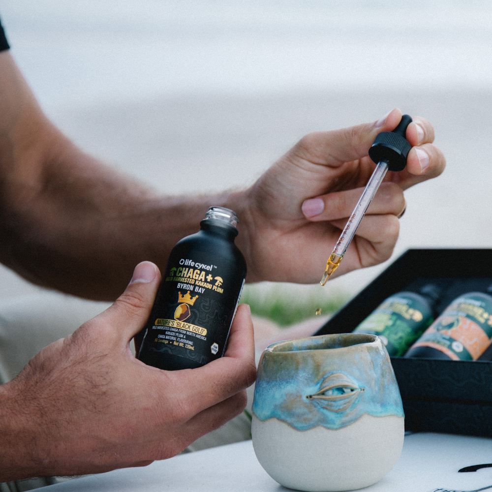 Person holding a Lifecykel Chaga mushroom extracts bottle and using a dropper with a ceramic cup on a table.