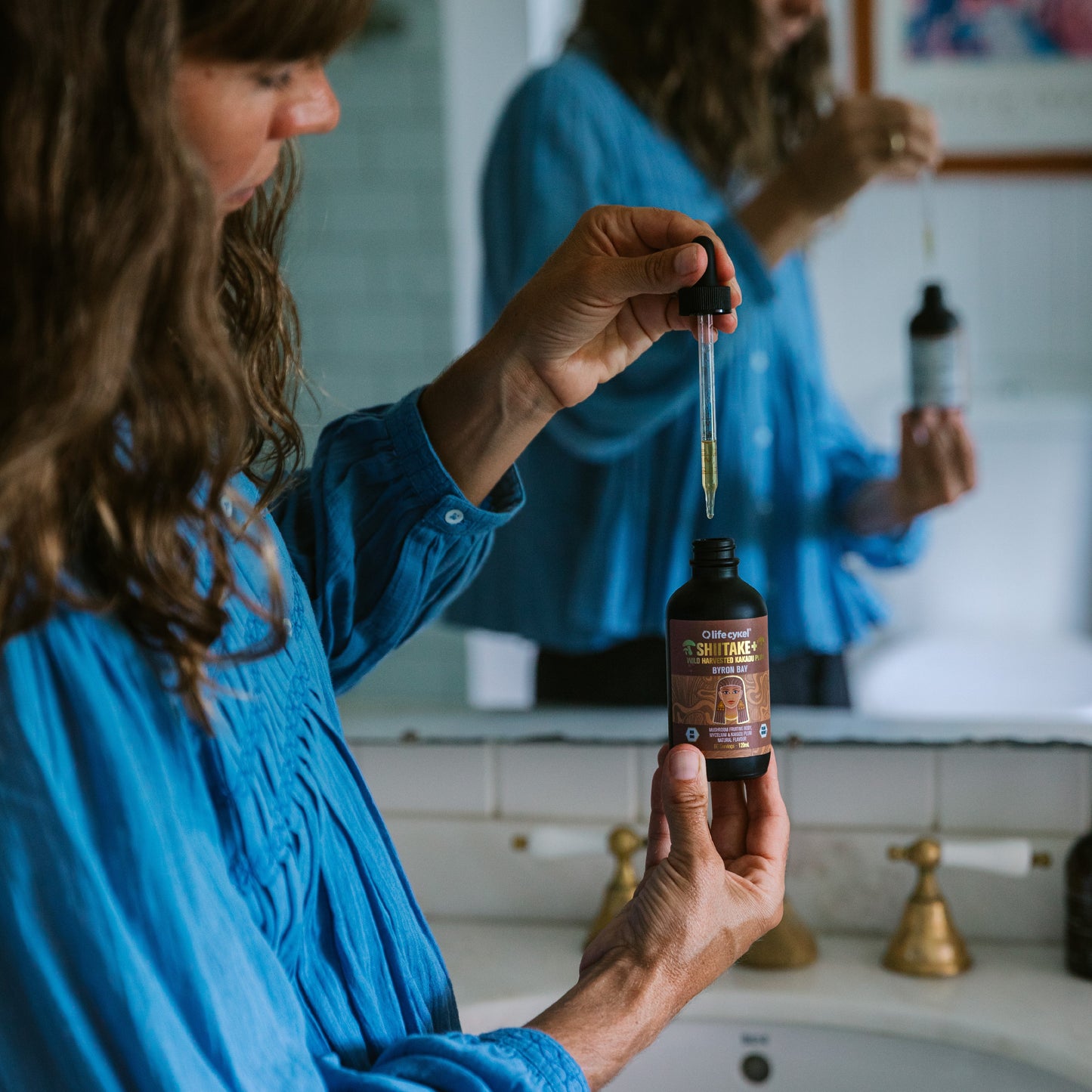 Woman in blue tops holding Lifecykel Reishi mushroom extracts with dropper in front of the mirror in the bathroom