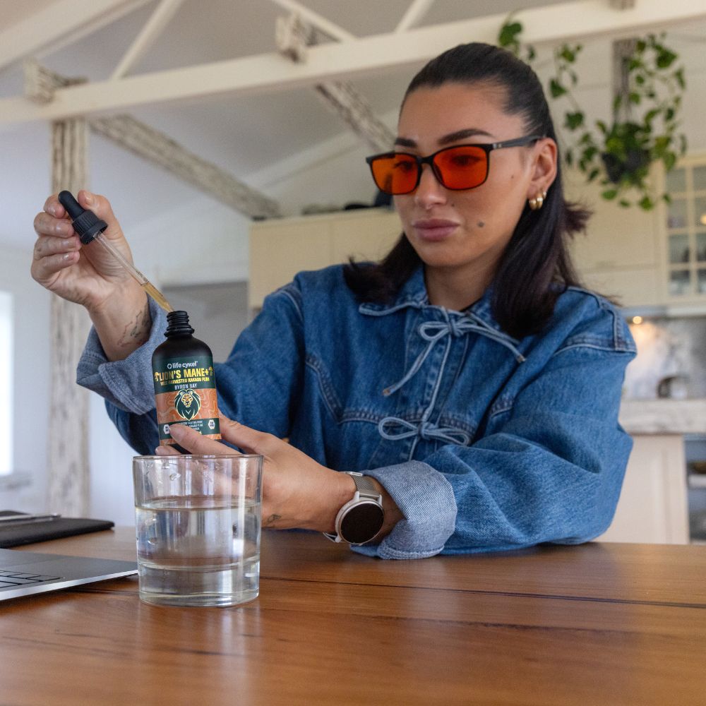 Person in a denim jacket and sunglasses pouring a Lifecykel Lion's Mane mushroom extracts liquid from a dropper bottle into a glass.
