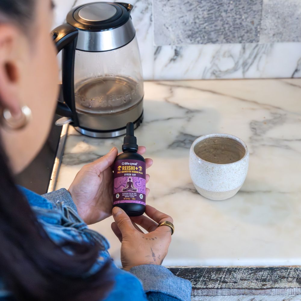 Person holding a bottle of 'Reishi' on a kitchen counter with a kettle and cup in the background.